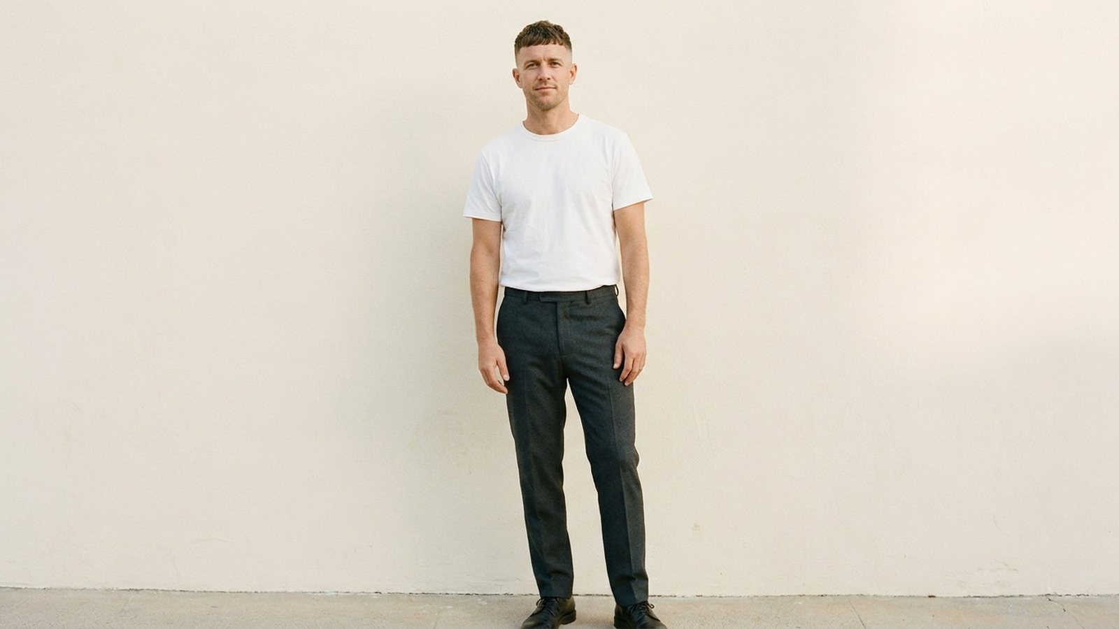 A male model standing against a plain studio wall in natural light